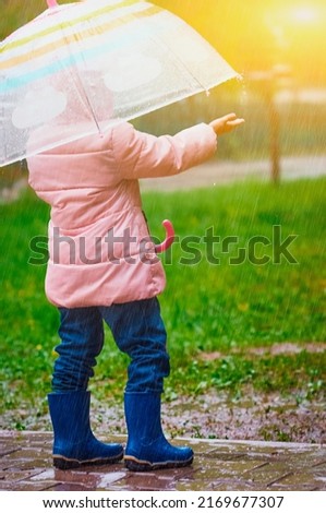 Rear view of a little girl in rubber boots with an umbrella in the pouring rain. Catches drops with the palm of your hand. Sunlight. Green grass is blurred in the background. Selective focus.