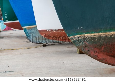 Two large fishing vessels on drydock in storage. One boat is blue with the number five in white. The second is green with the white lettering numbers eight, ten, and twelve. Both boats have red hulls
