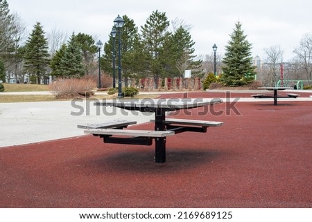 Wooden wheelchair accessible picnic table on a red pour in place rubber ground covering. There's a rollerblading skate park, tall pine trees and flower beds in the background under cloudy skies.