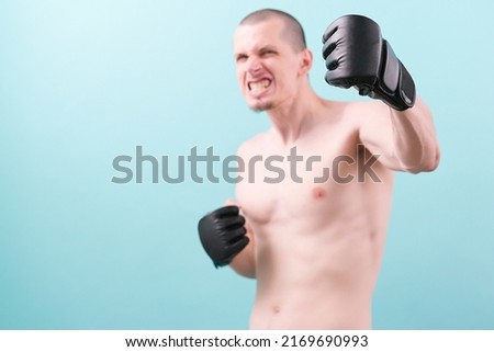 An angry MMA fighter holds a fist in black gloves on a blue background. Ready to make a punch. Teeth. Martial. Muscle. Workout. Aggressive. Man. Young. Competition. Guy. Handsome. Mixed. Muscular
