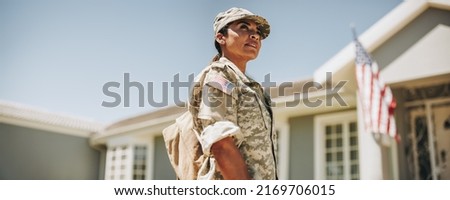 Courageous female soldier looking away thoughtfully while standing outside her house with her bag. American servicewoman coming back home after serving her country in the military. Royalty-Free Stock Photo #2169706015