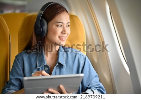 Happy and cheerful young asian woman with headphones using a portable tablet and looking the view outside of the plane window. Female passenger image Royalty-Free Stock Photo #2169709211