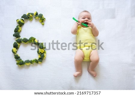 flatlay composition. Proper nutrition and complementary foods for babies concept. six-month-old baby plays with a green baby spoon, lies on a large bed next to the number six from green broccoli Royalty-Free Stock Photo #2169717357