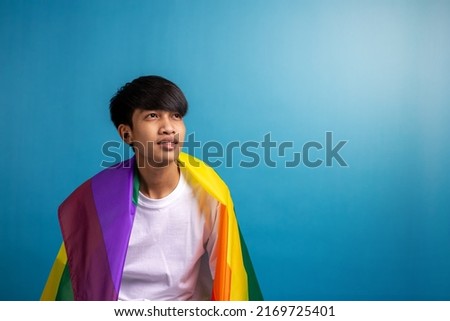 Asian young man with pride LGBT rainbow flag on shoulder against blue color background. A man with gay pride flag with copy space in studio.