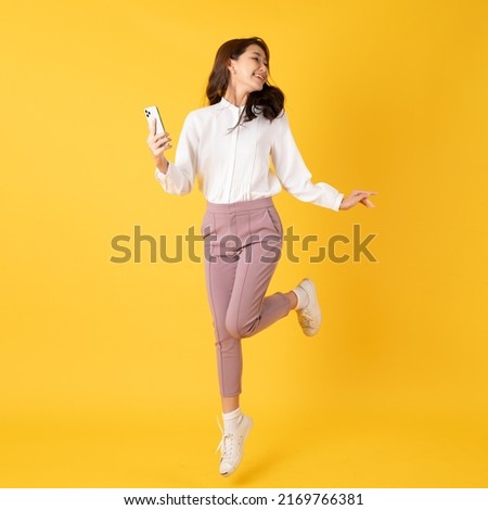 Smiling asian woman white shirt on yellow background jumping with smartphone on hand