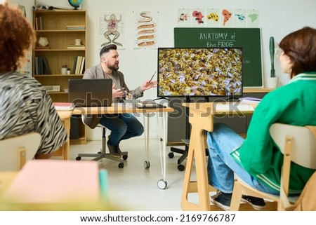 Confident mature teacher telling group of highschool students about life of bees while making presentation on interactive screen Royalty-Free Stock Photo #2169766787