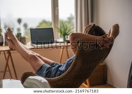 Relaxed woman in armchair with legs on table in front of open laptop in apartment