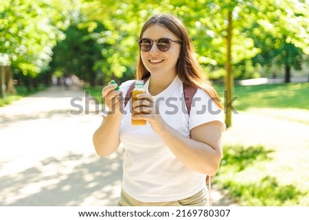 Young stylish happy woman with juice standing over nature background. Redhead girl walking in the park and holding a bottle of fresh cold smoothie in a warm sunny day.