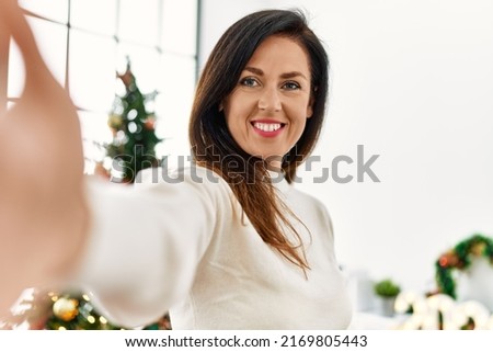 Middle age hispanic woman make selfie by the camera standing by christmas tree at home