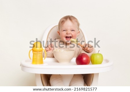 Baby wearing knitted sweater sitting in high chair and feels hungry, holding spoon and eating puree or porridge, enjoying apples and water in yellow bottle, posing isolated on white background. Royalty-Free Stock Photo #2169821577