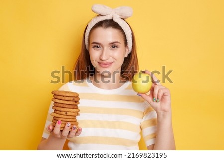 Portrait of happy young girl in hair band smiles broadly, cheerful woman in striped shirt holds fresh apple and tasty sweet cookie isolated on yellow background.