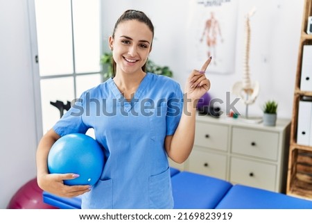 Young physiotherapist woman holding pilates ball at medical clinic smiling happy pointing with hand and finger to the side  Royalty-Free Stock Photo #2169823919