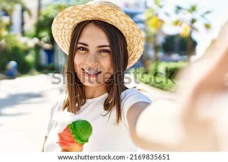 Young beautiful woman smiling happy outdoors on a sunny day wearing a summer hat and eating ice cream taking a selfie picture