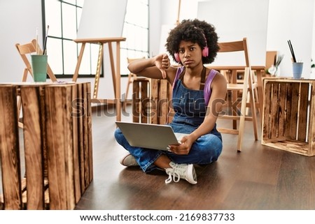 Young african american woman with afro hair at art studio using laptop with angry face, negative sign showing dislike with thumbs down, rejection concept 