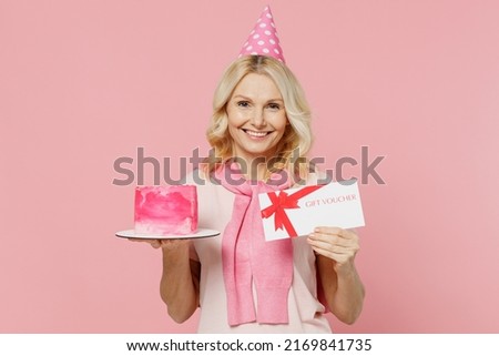 Elderly happy woman 50s wear t-shirt birthday hat hold cake gift certificate coupon voucher card for store isolated on plain pastel pink background studio portrait Celebration party holiday concept