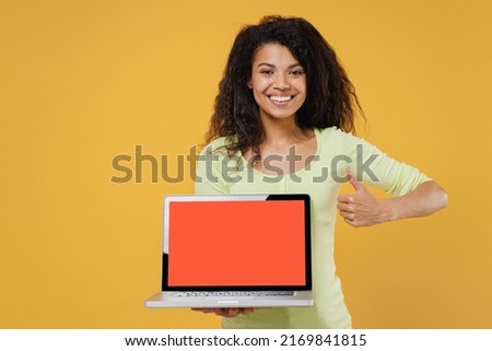 Fun african american young brunette woman 20s wears green shirt hold use work on laptop pc computer with blank screen workspace area show thumb up gesture isolated on yellow background studio portrait