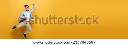Asian kid with backpack showing yes gesture and jumping on yellow background, banner Royalty-Free Stock Photo #2169843587