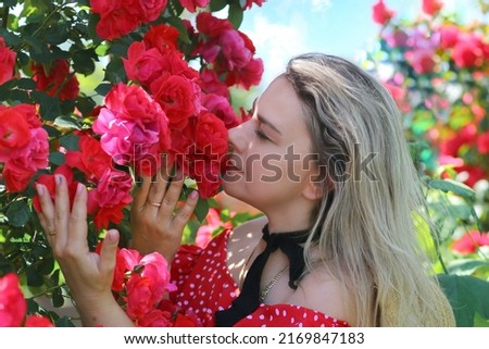 Flowers. Summer concept. Caucasian girl in a red dress with polka dots enjoying the scent of roses. Stunning flower garden backdrop