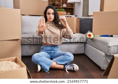 Young brunette woman sitting on the floor at new home holding keys with open hand doing stop sign with serious and confident expression, defense gesture 