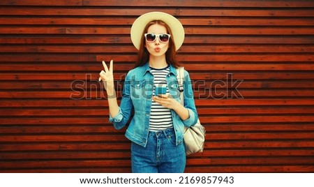 Portrait of young woman blowing her lips with phone wearing summer straw hat, backpack and jean jacket on wooden background
