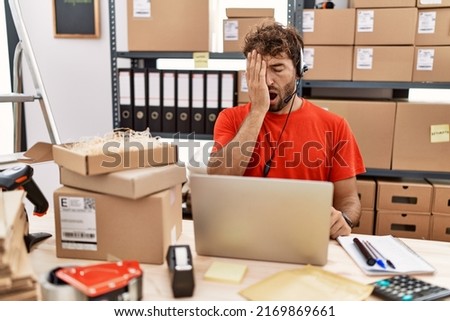 Young hispanic call center agent man working at warehouse yawning tired covering half face, eye and mouth with hand. face hurts in pain. 