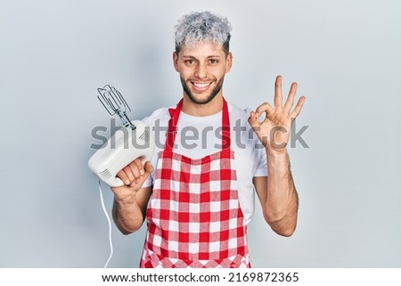 Young hispanic man with modern dyed hair holding food processor mixer machine doing ok sign with fingers, smiling friendly gesturing excellent symbol 
