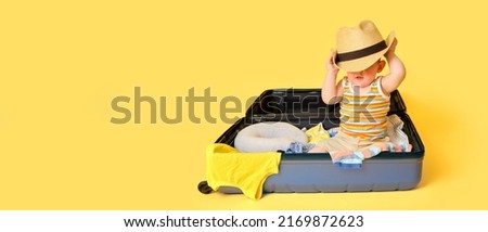 Baby toddler boy sits inside a suitcase with clothes, studio yellow background. A child plays in a big bag while packing luggage for a vacation trip, copy space. Kid age one year (12 months) Royalty-Free Stock Photo #2169872623