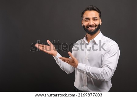 Indian male entrepreneur points with both hands aside isolated on black, bearded businessman presenting, showing novelty looking at the camera and smiles, copy space