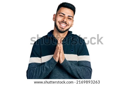 Young hispanic man with beard wearing casual winter sweater praying with hands together asking for forgiveness smiling confident. 