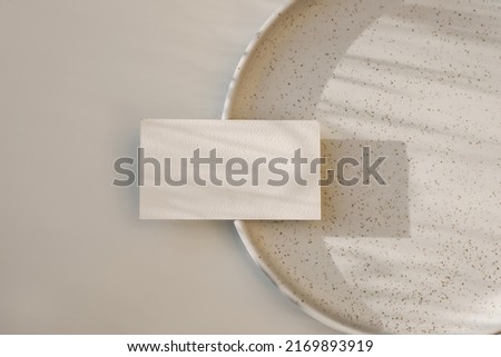 Modern neutral branding stationery. Closeup of textured blank business card, invitation mockup on dotted, speckled ceramic plate in sunlight. Beige table background. Flat lay, top view. No people.
