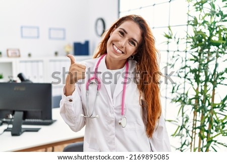Young redhead woman wearing doctor uniform and stethoscope at the clinic doing happy thumbs up gesture with hand. approving expression looking at the camera showing success. 