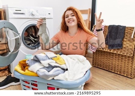 Young redhead woman putting dirty laundry into washing machine smiling looking to the camera showing fingers doing victory sign. number two. 