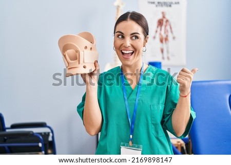 Young hispanic woman holding cervical neck collar at the clinic pointing thumb up to the side smiling happy with open mouth 