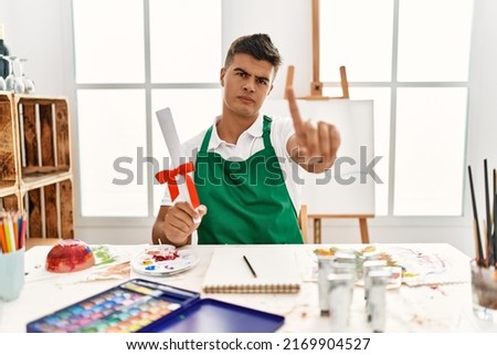 Young hispanic man at art studio holding degree pointing with finger up and angry expression, showing no gesture 