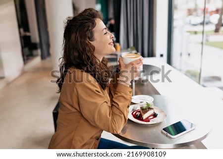 Profile picture of stylish European woman with curly hair wearing brown shirt enjoying coffee and breakfast in morning in modern cafeteria 