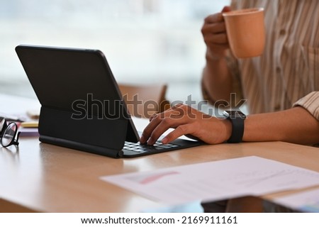 Cropped view businessman holding a cup of coffee sitting in an office and using a tablet, for business and technology concept.