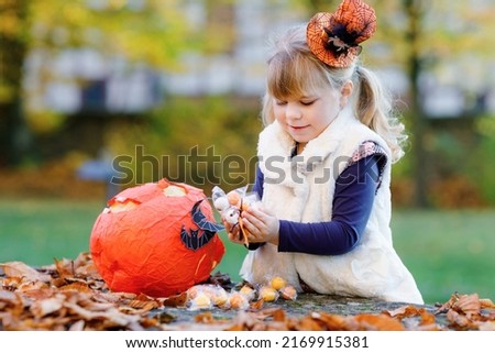 Little toddler girl dressed as a witch trick or treating on Halloween. Happy child outdoors, with orange funny hat and pumpkin bag for sweet haunt. Family festival season in october. Outdoor activity
