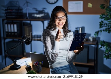 Young brazilian woman using touchpad at night working at the office doing money gesture with hands, asking for salary payment, millionaire business 