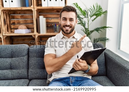 Handsome hispanic man holding clipboard working at psychology clinic cheerful with a smile on face pointing with hand and finger up to the side with happy and natural expression 