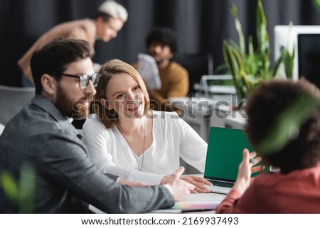 woman smiling near multiethnic colleagues and laptop with green screen in ad agency