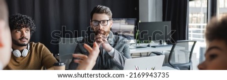 thoughtful man in eyeglasses near indian colleagues and blurred people in advertising agency, banner Royalty-Free Stock Photo #2169943735