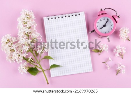 Notepad with a blank page for writing, alarm clock (clock) with an arrow at 7 o'clock, a sprig of blooming white lilac (syringa) on a pink background. Space for text. The concept of early awakening. G