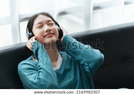 Beautiful asian woman in black headphones sits on sofa and enjoys listening to music, podcast.