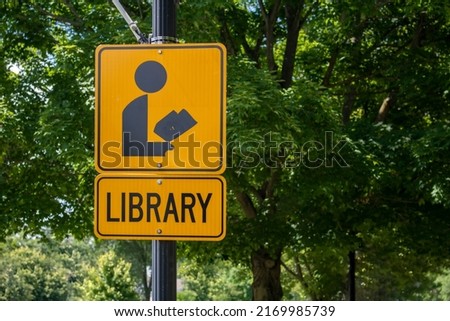 orange and black street and sidewalk sign with an icon of a figure reading a book with the word Library. Stating there's an of the public library nearby