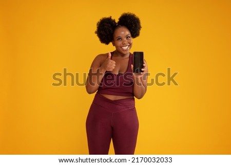 Smiling, happy, young girl with afro hairstyle posing on yellow studio background in sporty clothes, holding mobile phone in one hand showing new fitness app. Okay sign. Sale concept. Copy space. 