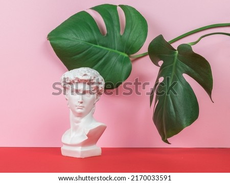 Plaster figure of Apollo on a red and pink background with large green leaves. Minimal concept. Creative photo.