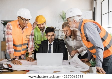 Four men and one woman sitting together at table and discussing blueprints on laptop during business conference. Multi ethnic architects, designers and engineers working at office.