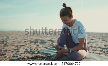 Beautiful woman sitting on yoga mat checking her smartphone on the beach. Young yogi woman looking happy resting with mobile phone by the sea