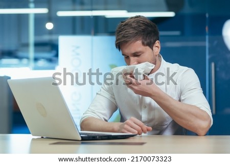 Feeling unwell in the workplace. Young handsome male businessman, freelancer getting sick, wiping his nose with a napkin, flu