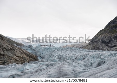 Jostedalsbreen Glacier in Norway forms a rough landscape of cracks and crevasses in the valley between a rocky mountain range. With blue, white, and grey ice. Picture for adventure tour advertisement.
